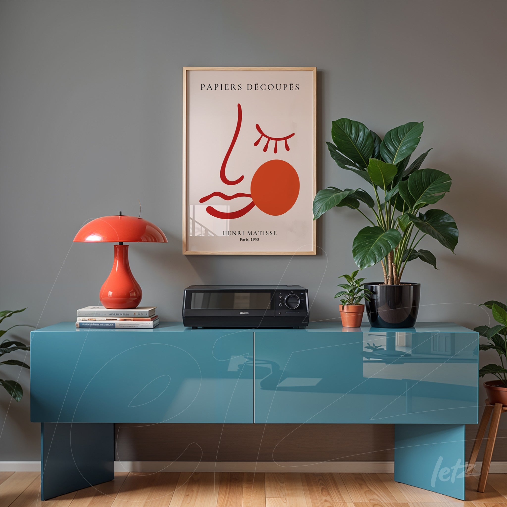 framed modern art of a stylized face in gold frame displayed above a blue table with plants and red lamp