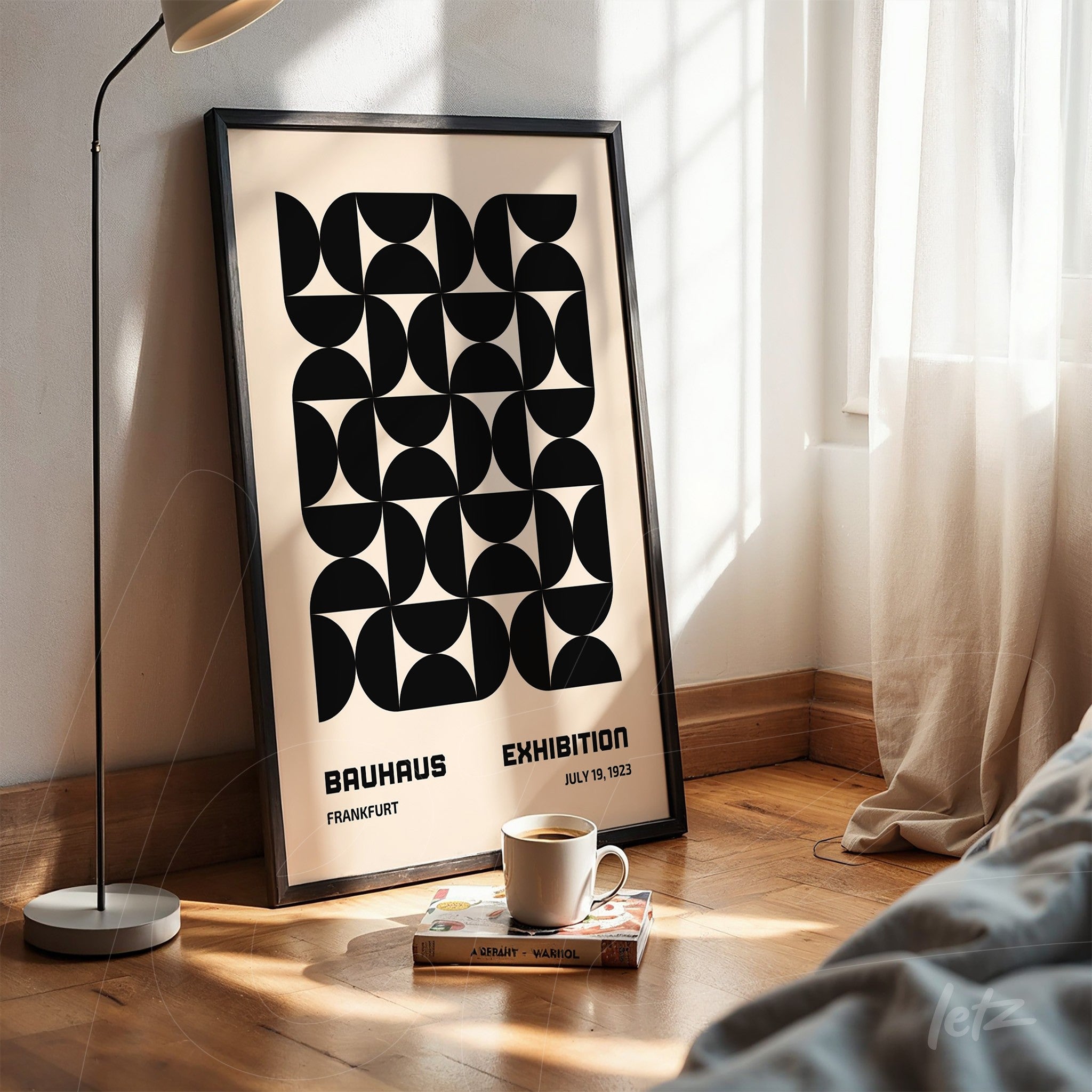 poster featuring black geometric design on beige background in black frame leaning against a white bedroom wall with lamp and cup on the floor