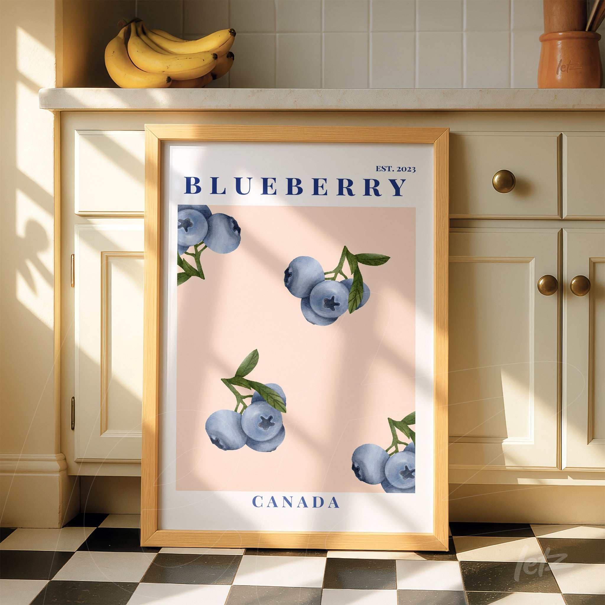 framed wall art featuring blueberries on a light pink background with a light wooden frame displayed in a bright kitchen