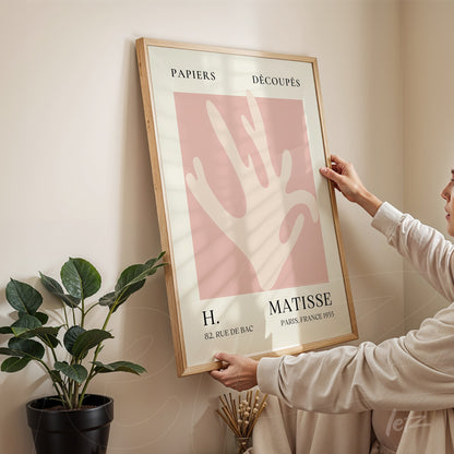 framed wall art featuring abstract shapes in pink and beige, light wood frame displayed against a light wall with a plant in the background