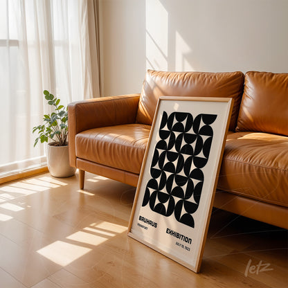 poster print featuring black geometric design in light wood frame resting on brown leather sofa in a sunlit room