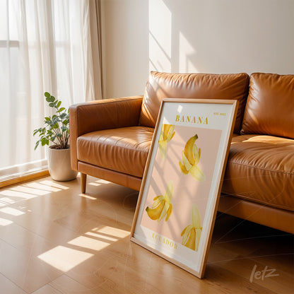 framed artwork featuring bananas on light pink background leaning next to a brown sofa in a sunlit room