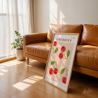 decorative wall art of illustrated cherries in white frame leaning against light brown sofa with natural light
