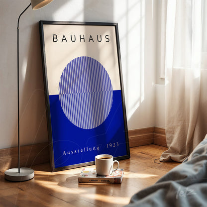 bauhaus poster print with blue sphere and white lines in black frame, resting on wooden floor next to coffee cup