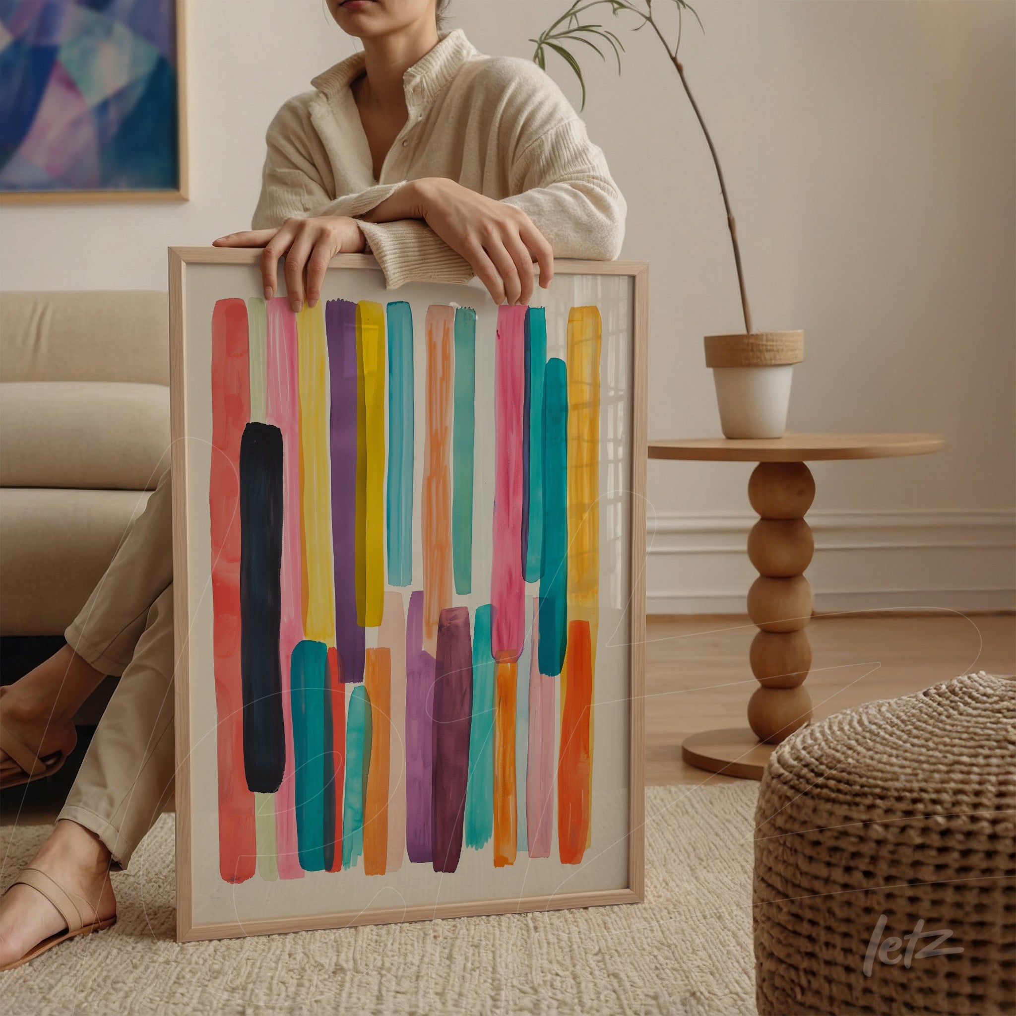 framed wall art featuring abstract color stripes in vibrant shades of blue, pink, yellow, and purple, leaning against a round wooden table next to a seated woman