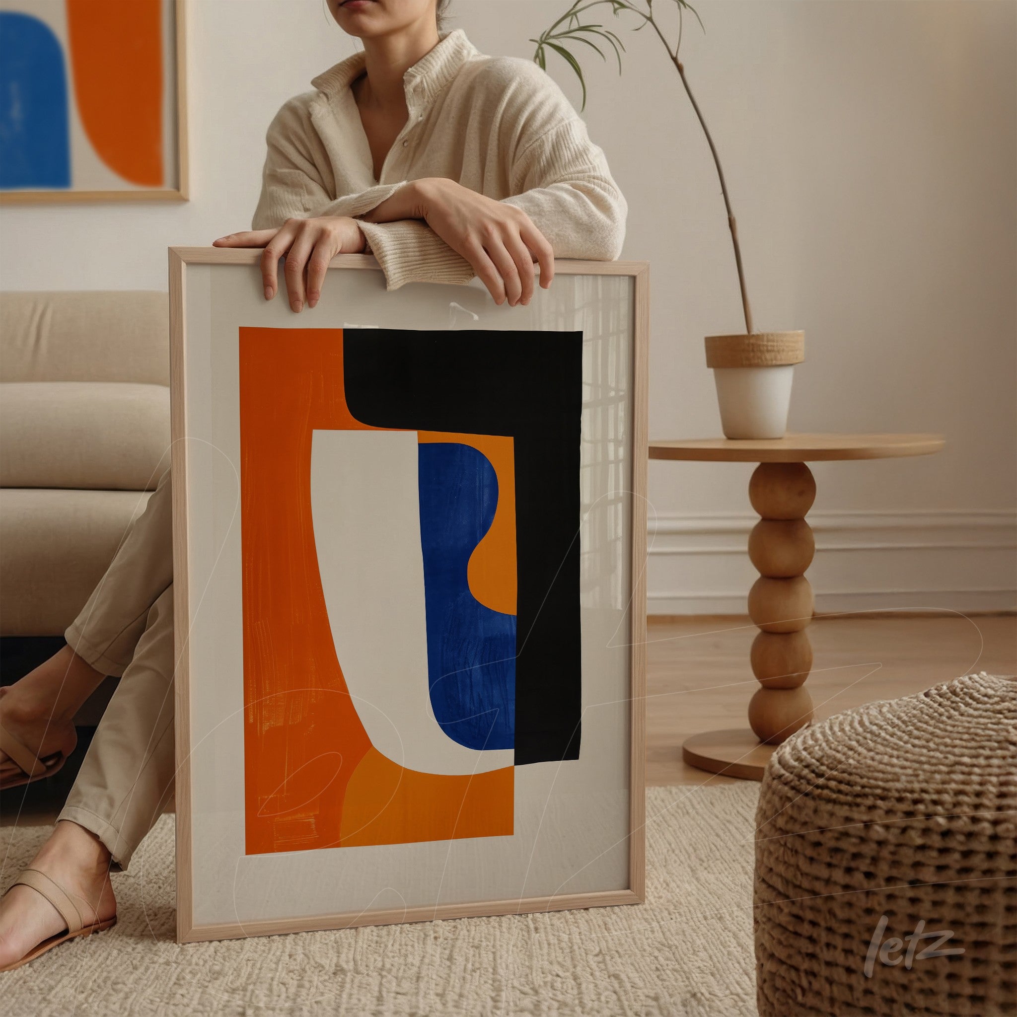 modern artwork featuring abstract shapes in orange, white, and black, framed in light wood, positioned next to a round side table